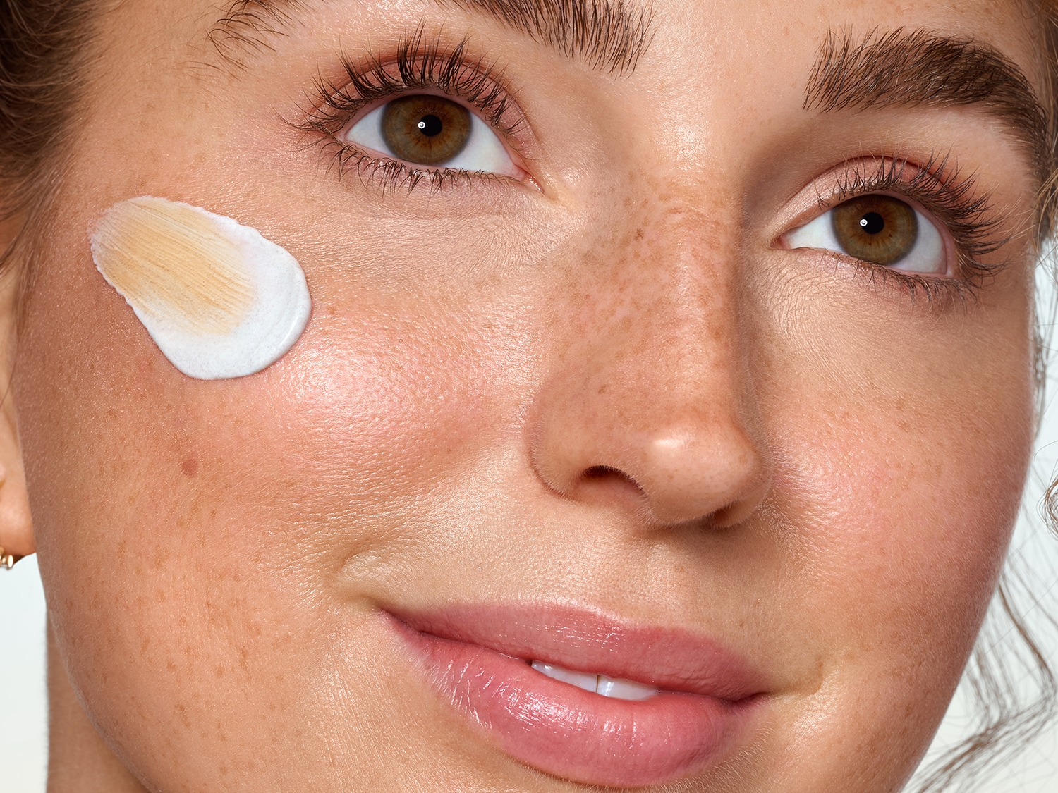 Gros plan sur le visage d'une femme avec une touche de crème blanche et beige sur la joue, mettant en valeur sa peau claire et ses taches de rousseur.