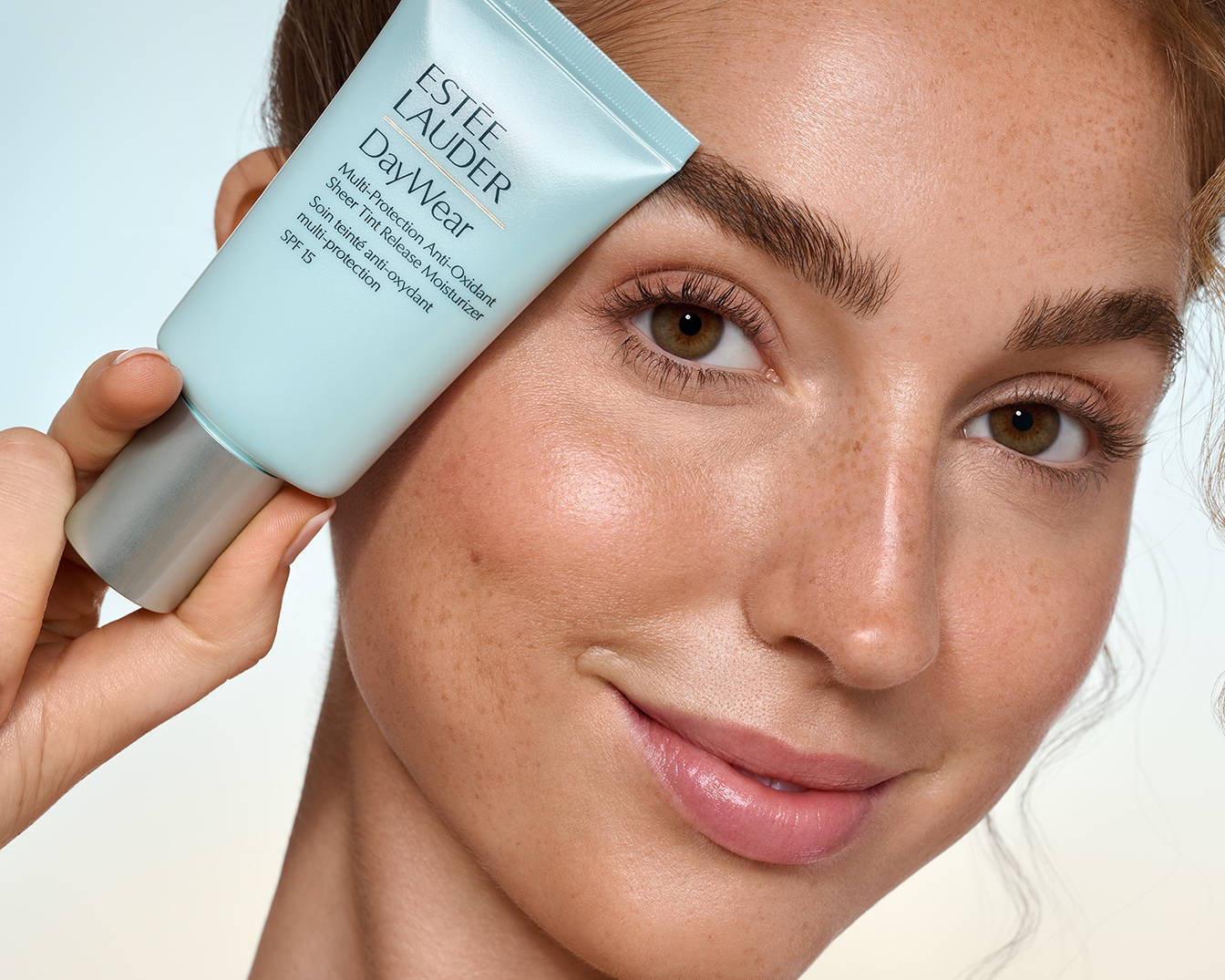 Une femme tient un tube bleu clair d'Estée Lauder DayWear Multi-Protection Anti-Oxidant Sheer Tint Release Moisturizer SPF 15, mettant en valeur son teint éclatant et sa peau protégée.