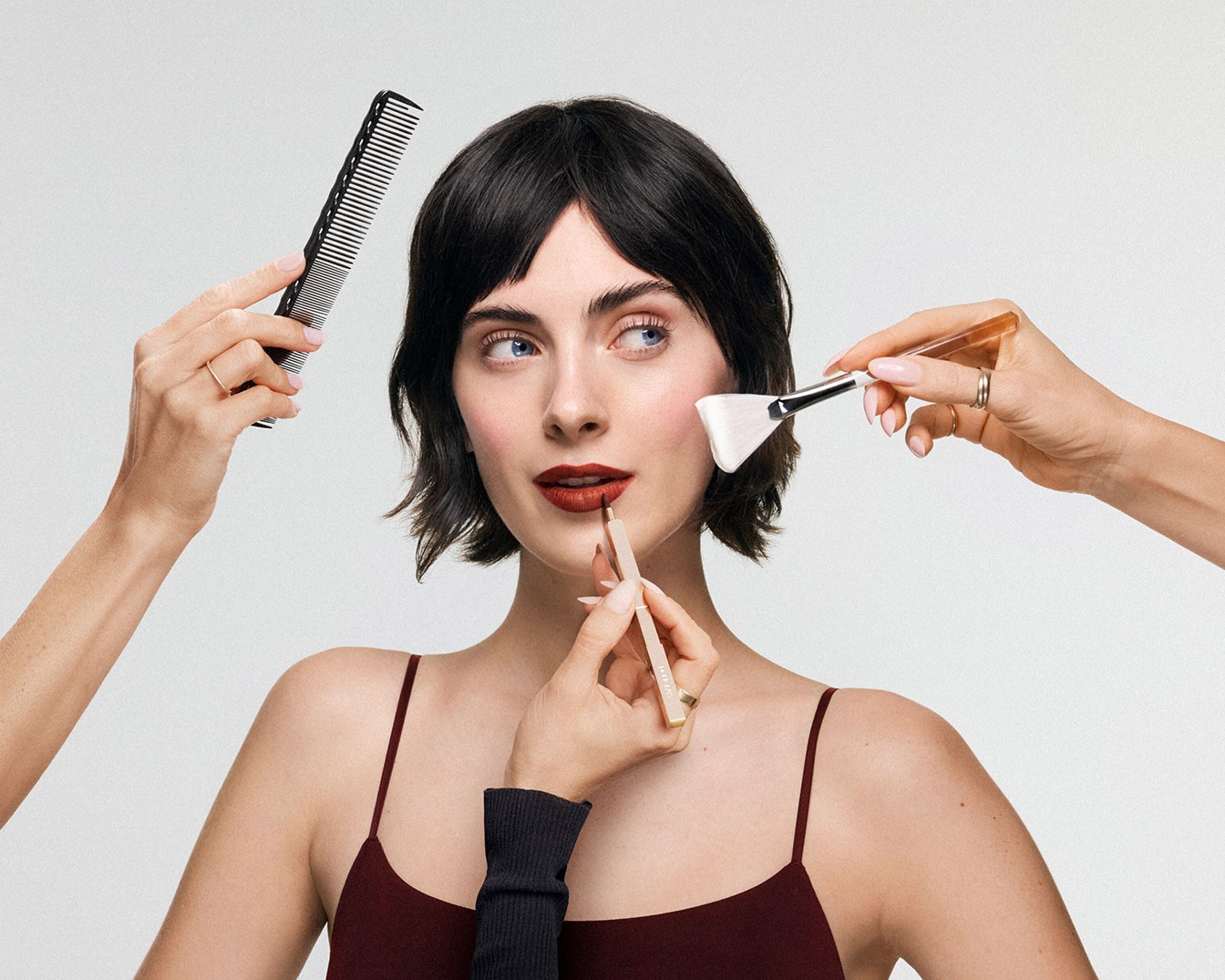 Une femme aux cheveux bruns se fait maquiller les lèvres avec un crayon à lèvres marron, tandis qu'une main tient un peigne noir et une autre un pinceau à maquillage.