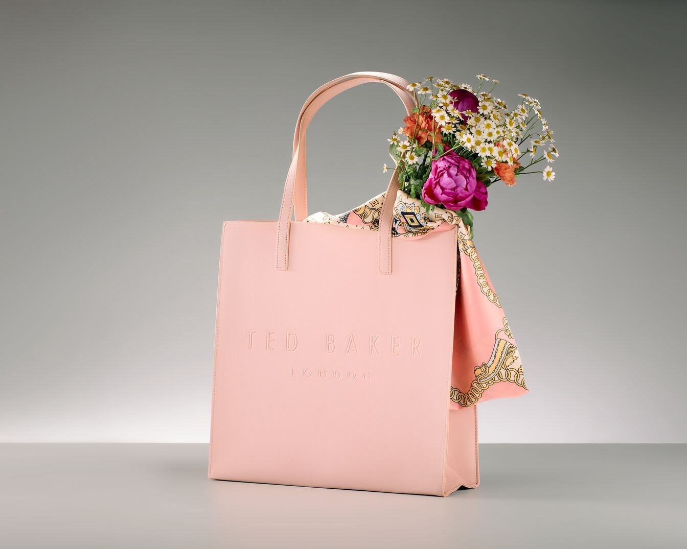 Nahaufnahme einer hellrosa Ted Baker Handtasche mit Henkeln, gefüllt mit einem bunten Blumenstrauß und einem gemusterten Tuch, vor einem neutralen Hintergrund.