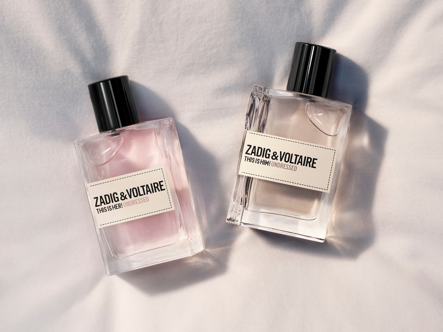 Dos frascos de perfume Zadig & Voltaire "This is Her! Undressed" con un líquido rosado y "This is Him! Undressed" con un líquido color crema, ambos con tapas negras sobre una tela blanca.