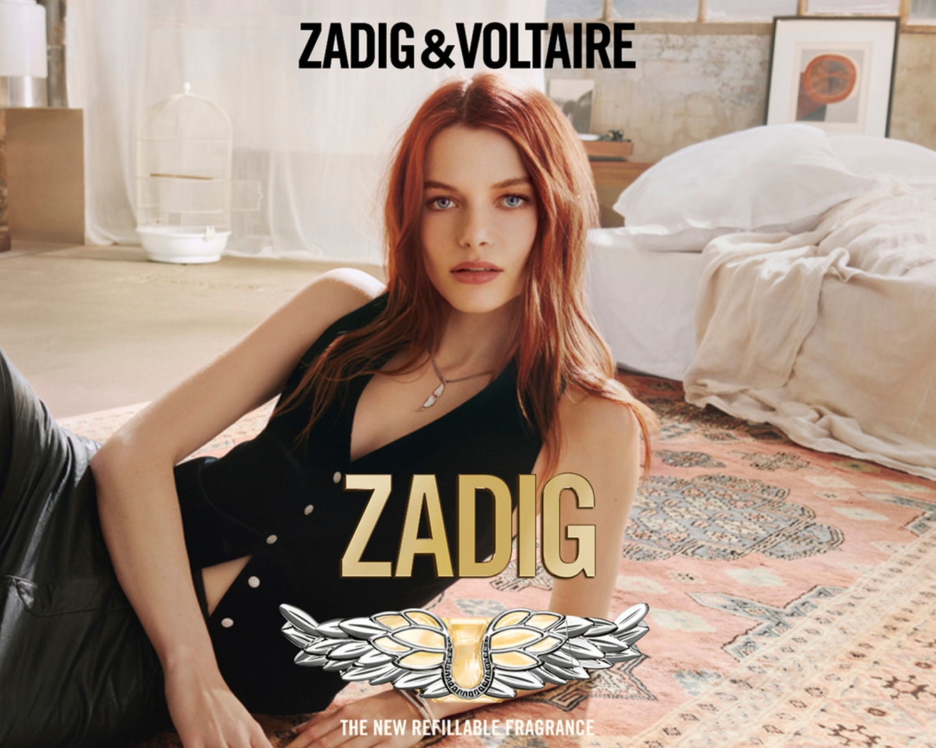 Una mujer pelirroja con ojos azules yace sobre una alfombra estampada con un chaleco negro, promocionando la nueva fragancia recargable de Zadig & Voltaire.