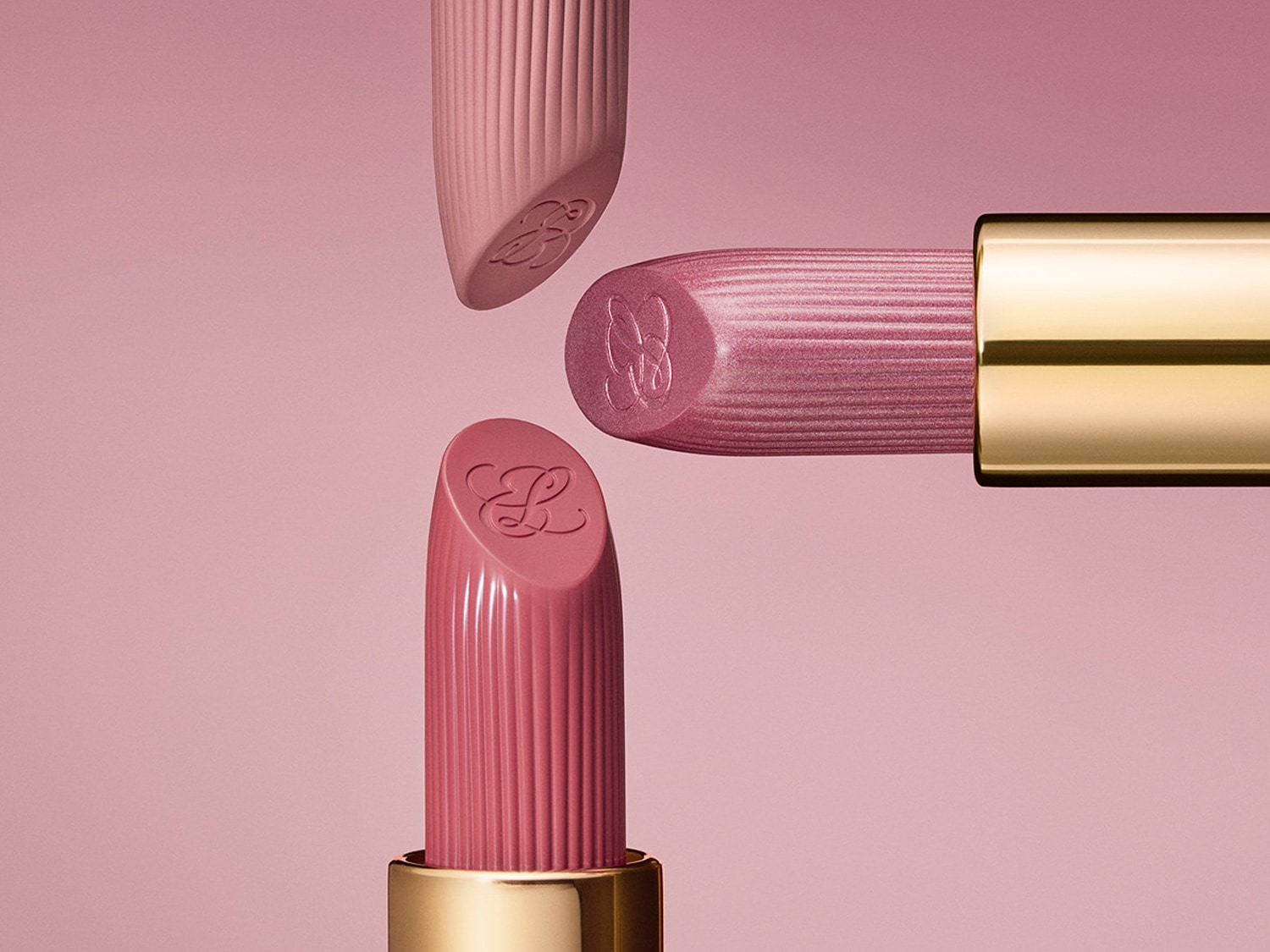 Trois rouges à lèvres rose pastel avec des rainures délicates et un logo en relief, présentés sur un fond rose assorti pour une élégance subtile.