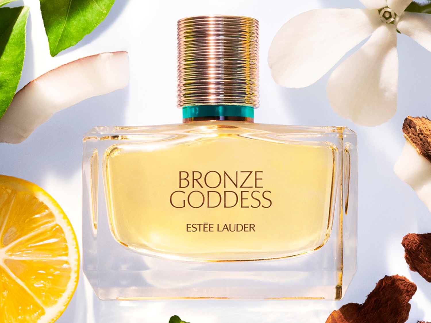Le parfum Bronze Goddess d'Estée Lauder dans un flacon transparent, entouré de citron, de noix de coco et de fleurs blanches.