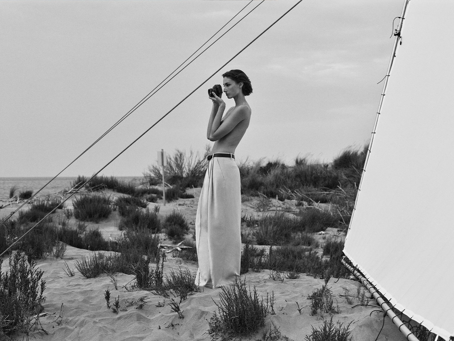 Čiernobiela fotografia ženy, ktorá stojí na pláži a drží fotoaparát pri tvári, oblečená v širokých nohaviciach s opaskom.