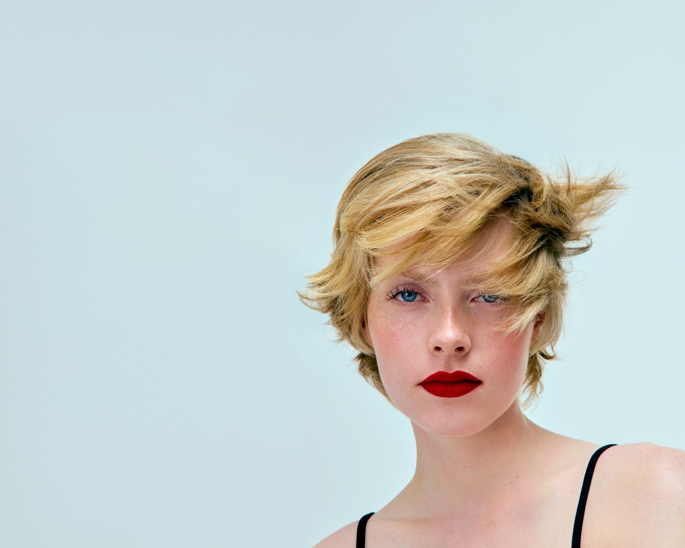 Portrait d'une femme aux cheveux blonds courts et aux lèvres rouge vif, sur un fond bleu clair, mettant en valeur un look audacieux et moderne.