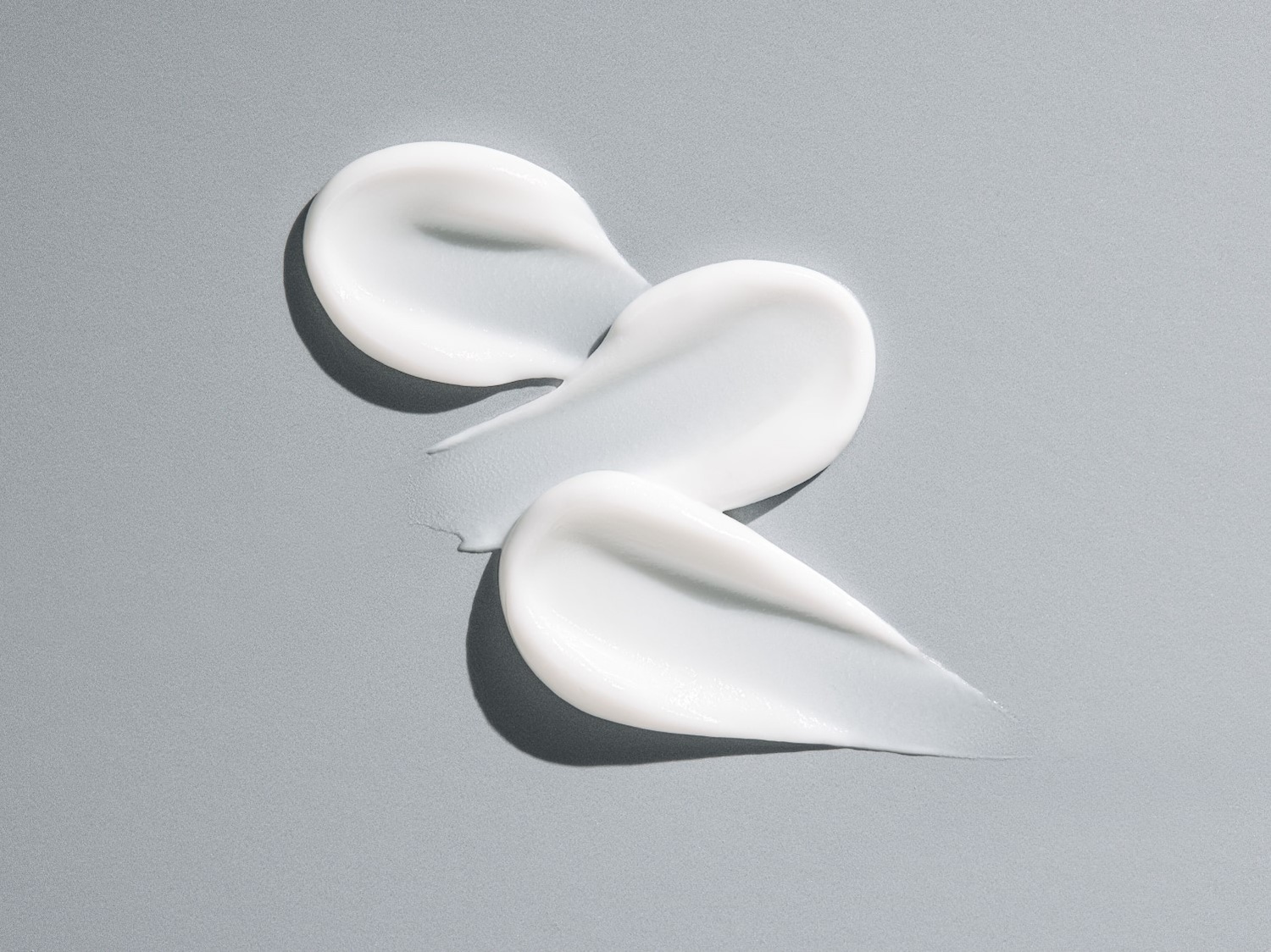 Drie vegen witte crème op een grijze achtergrond, die de gladde textuur en hydraterende eigenschappen van het product benadrukken.