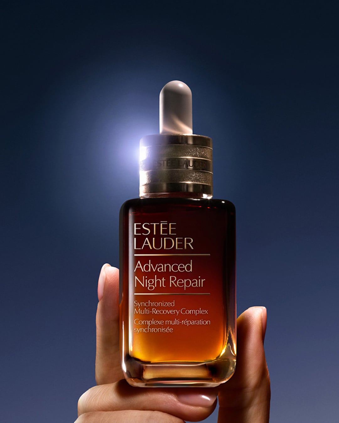 Een hand houdt een flesje Estée Lauder Advanced Night Repair vast, met een gouden vloeistof en een witte applicator, tegen een donkerblauwe achtergrond.