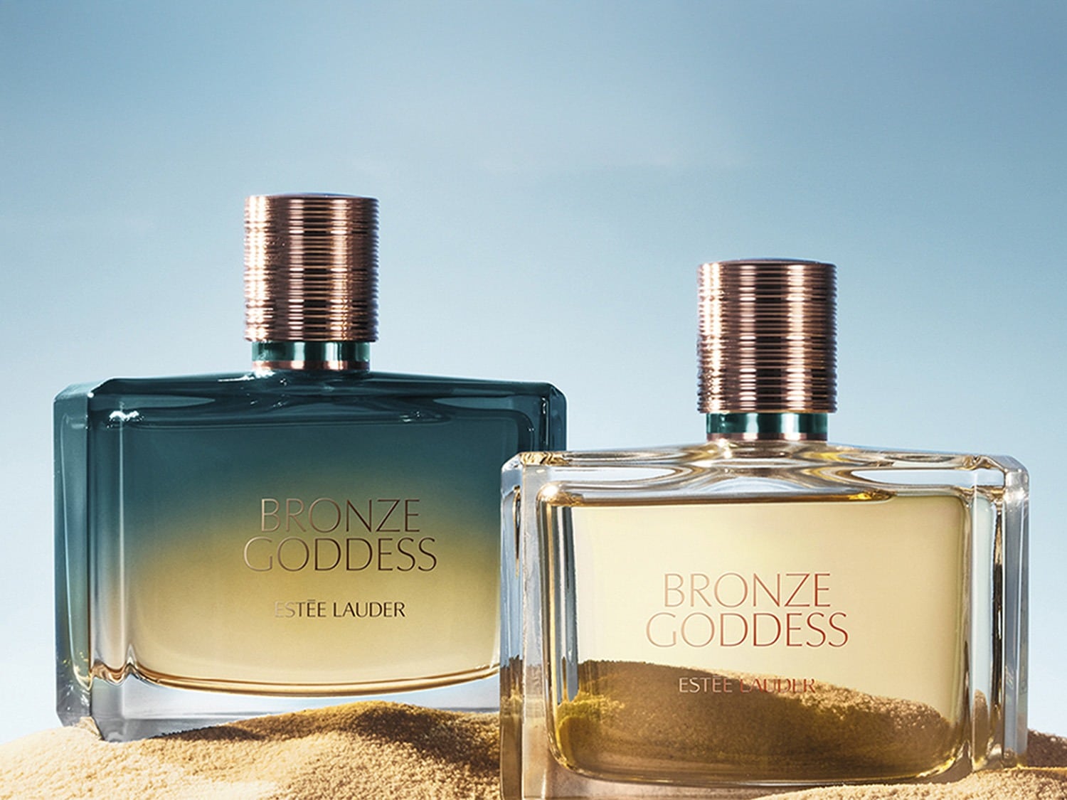 Twee flessen Estée Lauder Bronze Goddess parfum staan op een zandkleurige ondergrond, de ene fles heeft een blauw-gouden kleurverloop, de andere is helder met een gouden tint.