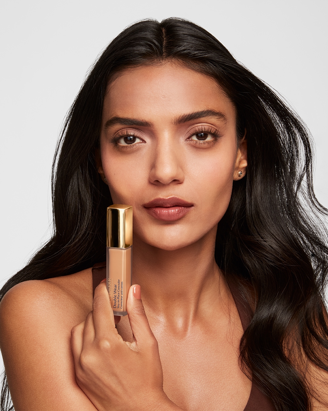 Een vrouw met een getinte huid houdt Estée Lauder Double Wear concealer vast, de verpakking is goudkleurig en de vloeistof is beige.