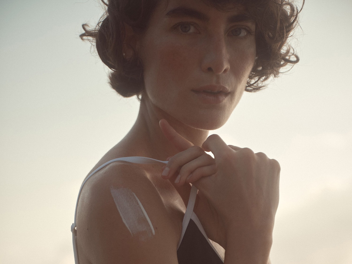 Una mujer de piel clara con el pelo castaño rizado se aplica crema solar en el hombro, preparándose para disfrutar del sol con protección.