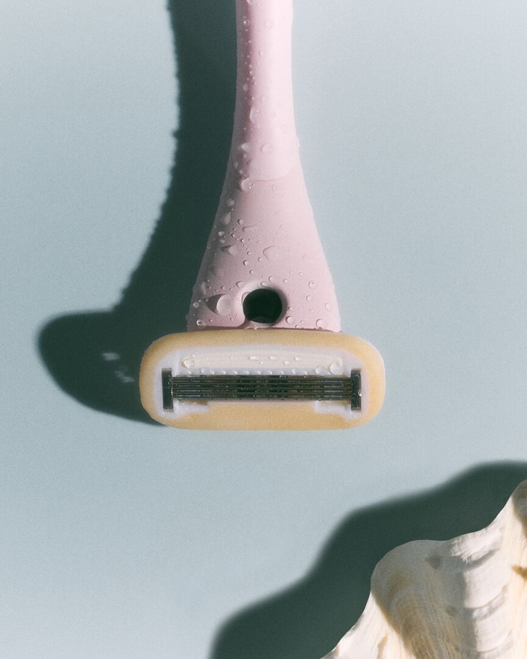 Primer plano de una maquinilla de afeitar rosa con gotas de agua sobre un fondo azul claro, con una concha blanca en la esquina inferior derecha.