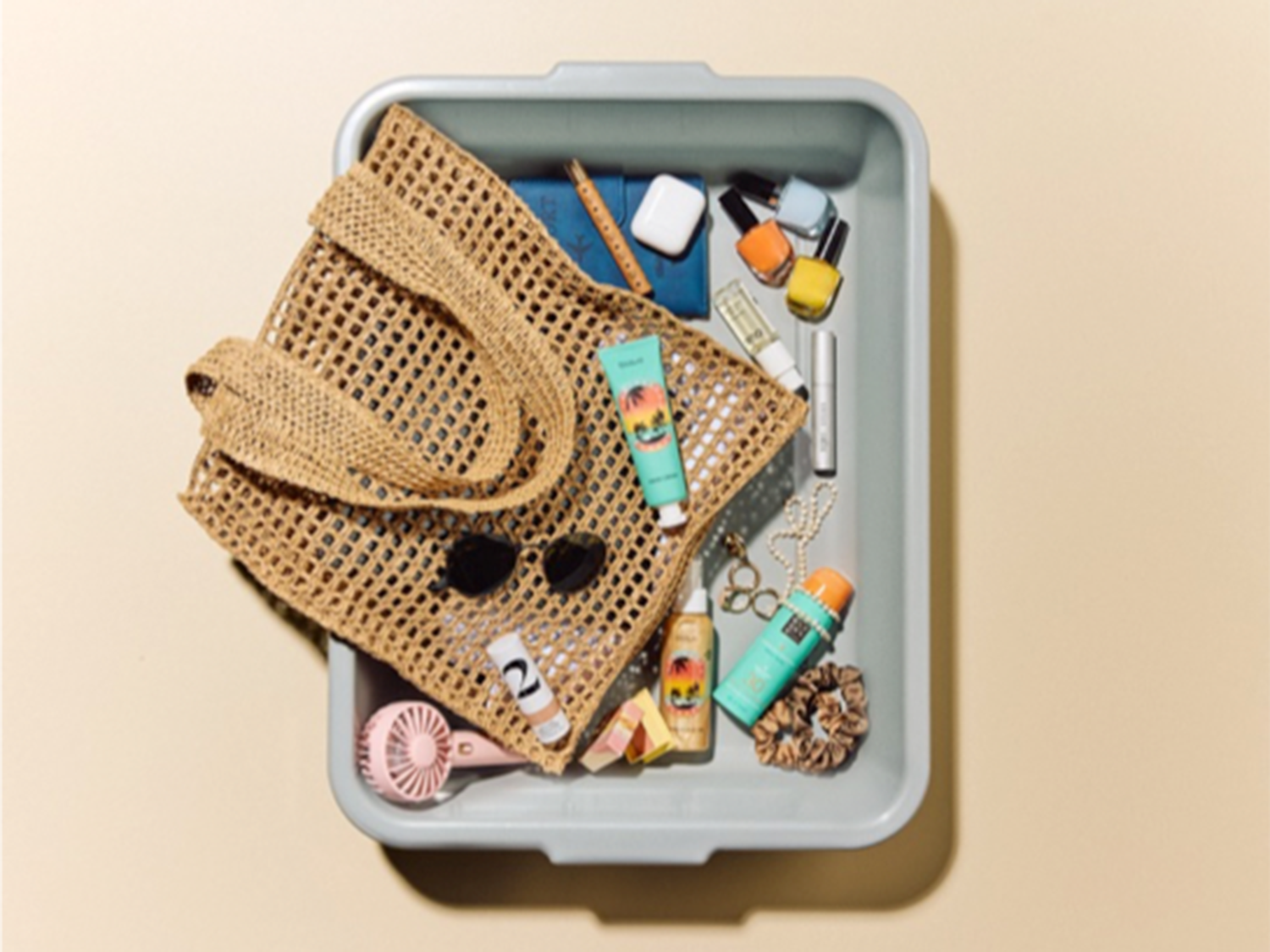 En una bandeja gris, un bolso de mano de rejilla marrón contiene gafas de sol, un mini ventilador rosa, cosméticos y accesorios, listos para un día de playa.