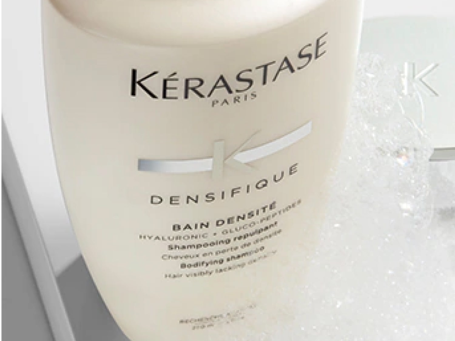 Nahaufnahme der hellen, cremefarbenen Flasche Kérastase Paris Densifique Bain Densité Shampoo, das mit Hyaluronsäure und Gluco-Peptiden formuliert ist, um das Haar sichtbar zu verdichten.