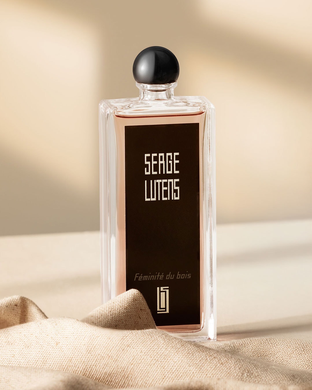 Ein transparenter Flakon von Serge Lutens "Féminité du bois" mit einem schwarzen Etikett und Verschluss, der auf einem beigen Stoff drapiert ist.