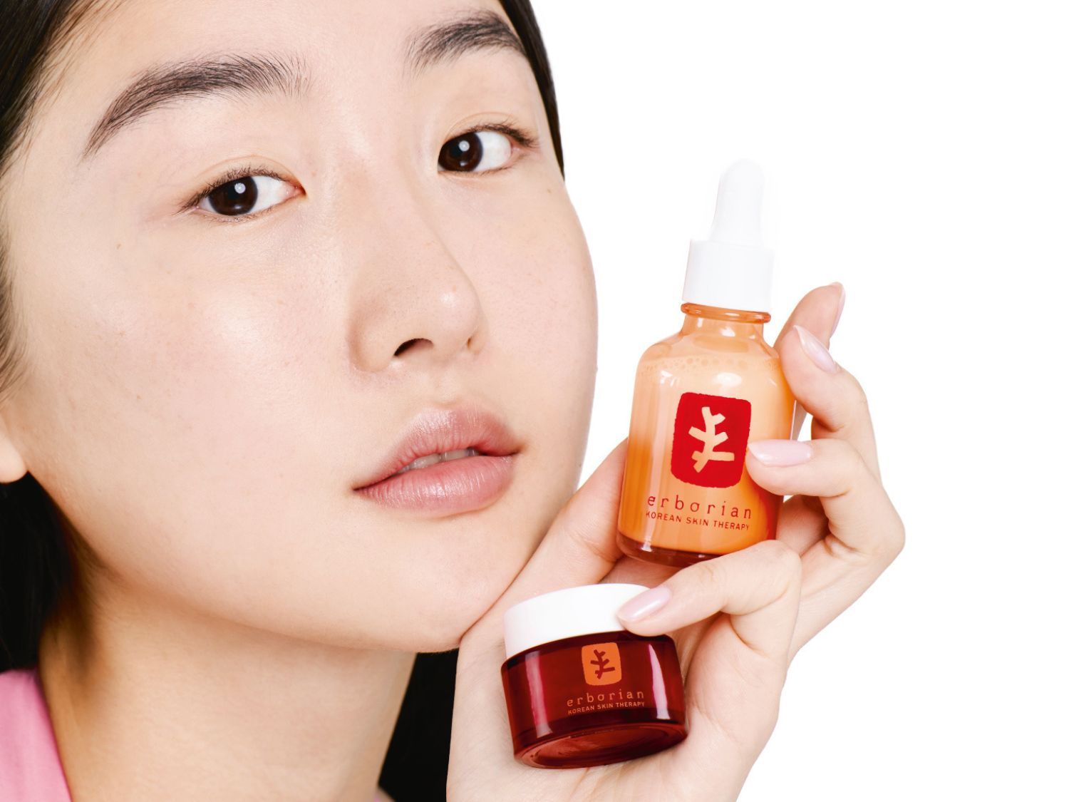 Arcához közel tartva, egy nő az Erborian koreai bőrápoló termékeit mutatja be: egy narancssárga üvegcsét fehér pipettával és egy piros tégelyt, mindkettőn a márka logója látható.