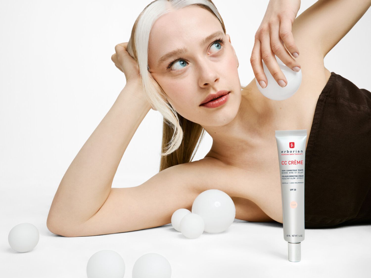 Na obrázku je žena s blond vlasmi a modrými očami, ktorá leží vedľa bielej fľaštičky CC krému Erborian s SPF 30 a sľubom zdravej a žiarivej pleti.