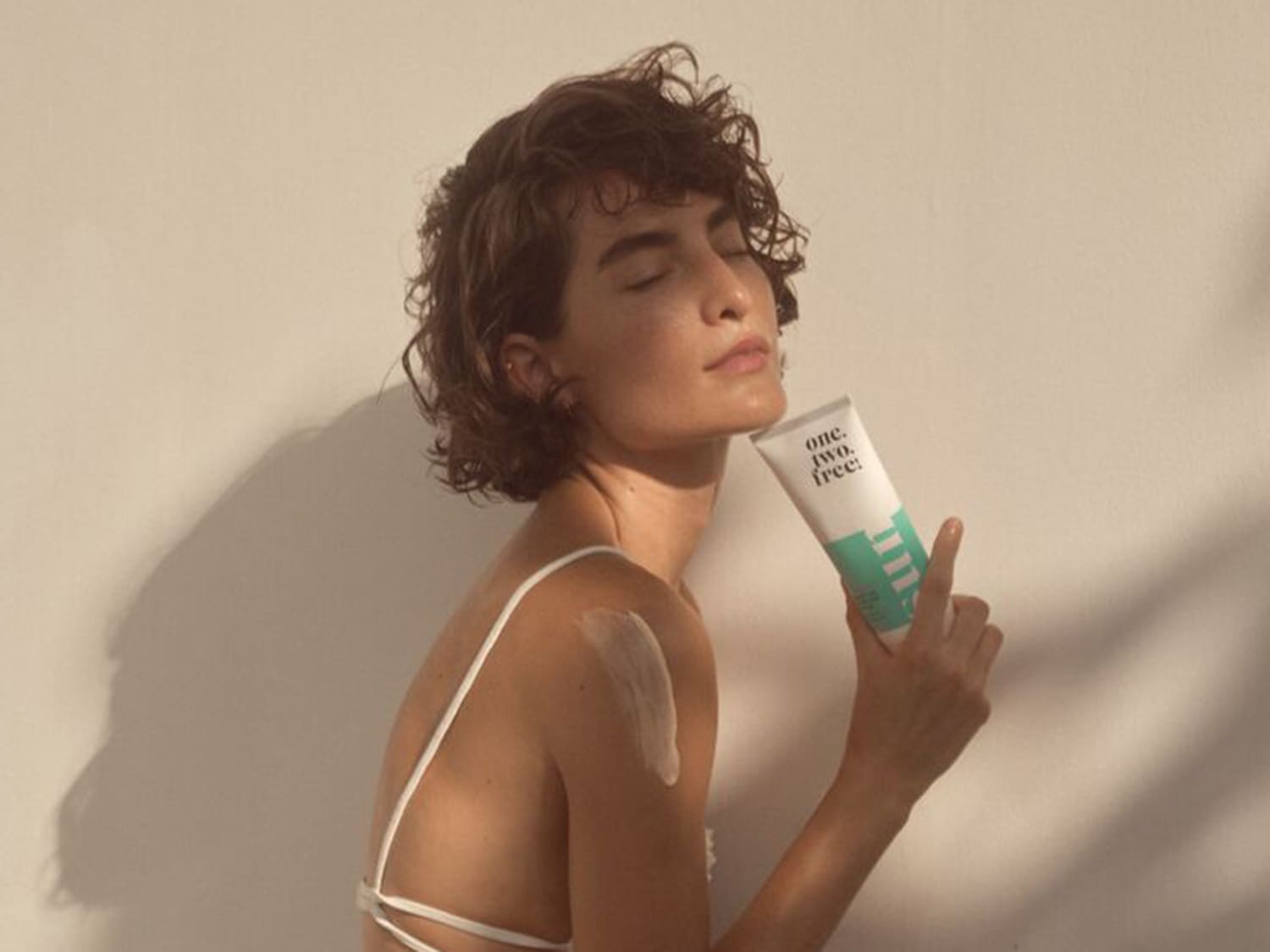 Una mujer de piel clara con el pelo corto y rizado sostiene un tubo blanco y verde de crema hidratante One. Two. Free, con una mancha de crema en el hombro.