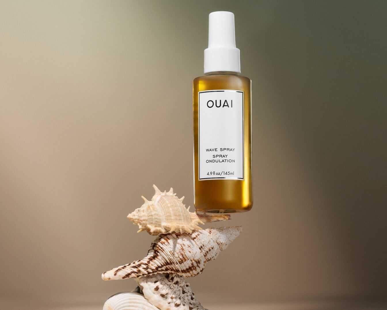 El spray para ondas OUAI de 4.9 fl oz/145 ml está sobre una pila de conchas marinas, ofreciendo una solución para crear ondas playeras sin esfuerzo.