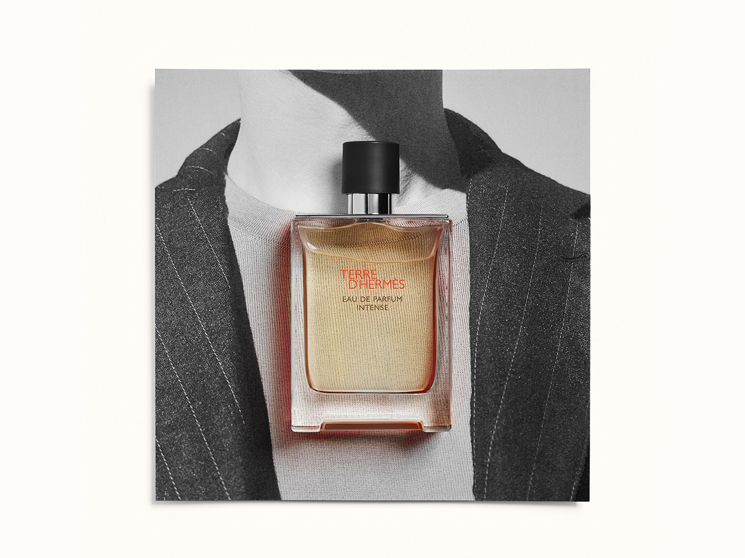 El frasco transparente de Terre d'Hermès Eau de Parfum Intense revela un líquido ámbar, resaltando su elegancia sobre un fondo de traje a rayas en blanco y negro.