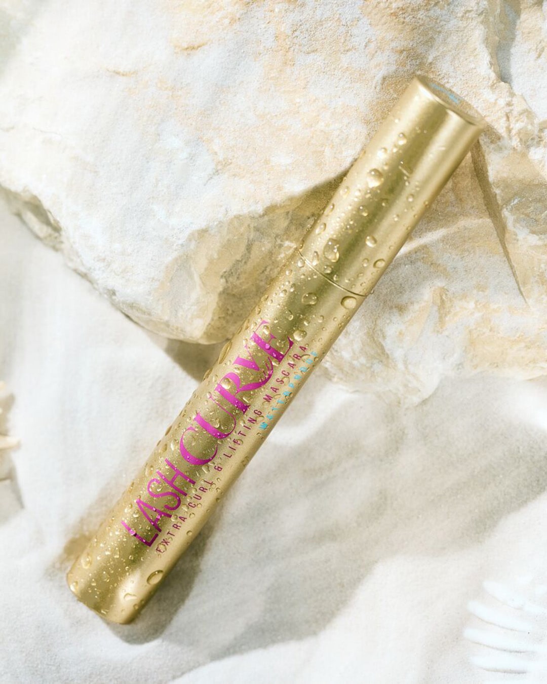 Auksinės spalvos tušas blakstienoms „Lash Curve Extra Curl & Lifting Mascara“ su rožiniu užrašu, padengtas vandens lašeliais, guli ant smėlio šalia akmens.