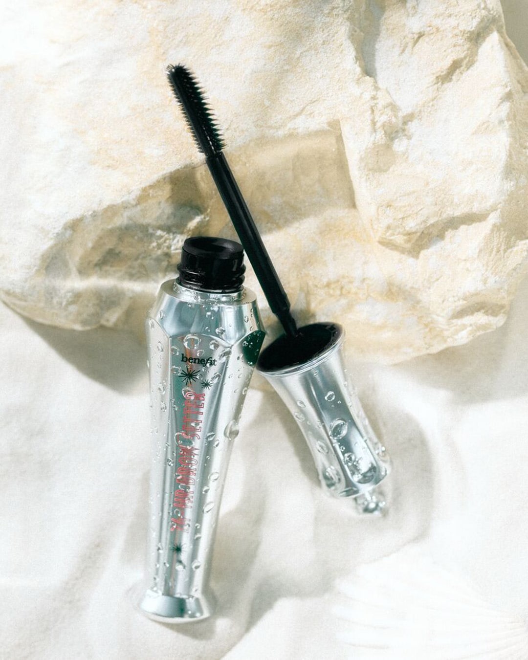 Sidabrinės spalvos „Benefit Fluff Up Brow Setter“ antakių gelio buteliukas su juodu šepetėliu guli ant smėlio šalia akmens.