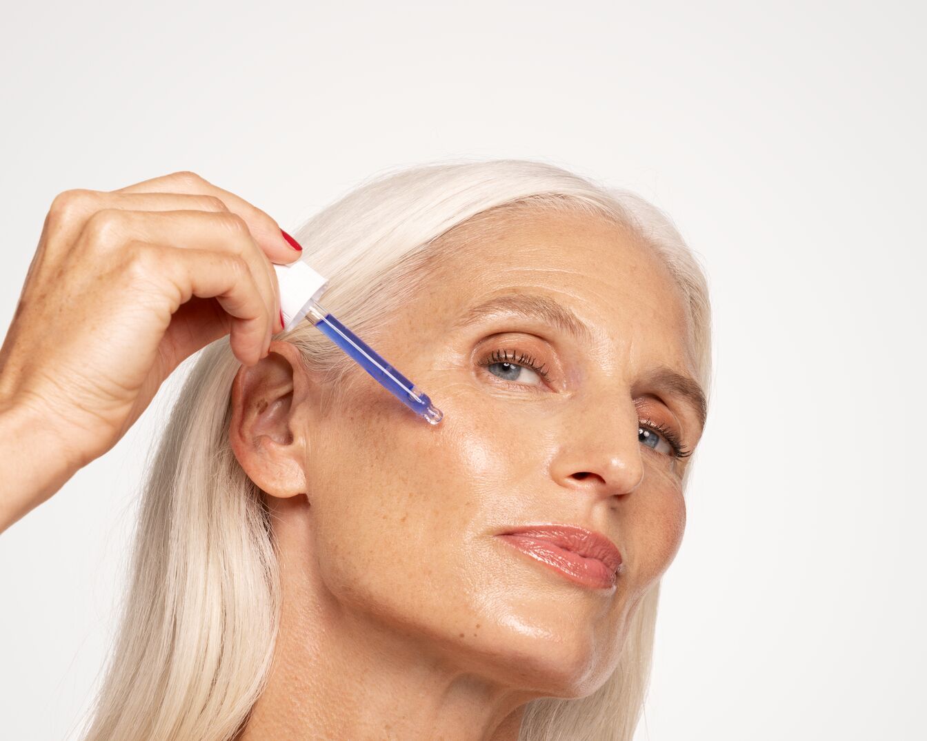 Eine Frau mit grauen Haaren trägt ein blaues Serum mit einer Pipette auf ihr Gesicht auf, um ihre Haut zu pflegen.