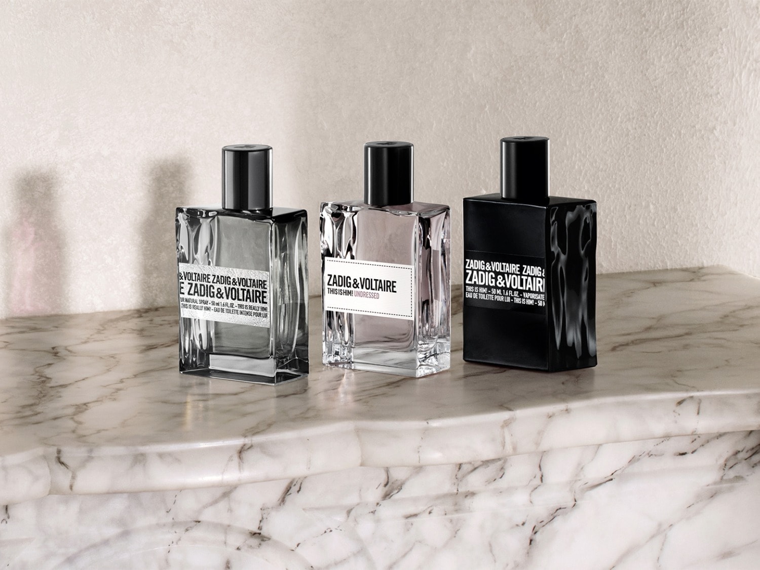 Trois flacons de parfum Zadig&Voltaire sont présentés sur une surface en marbre, chacun avec un design et une couleur distincts, offrant un aperçu de la gamme de parfums.