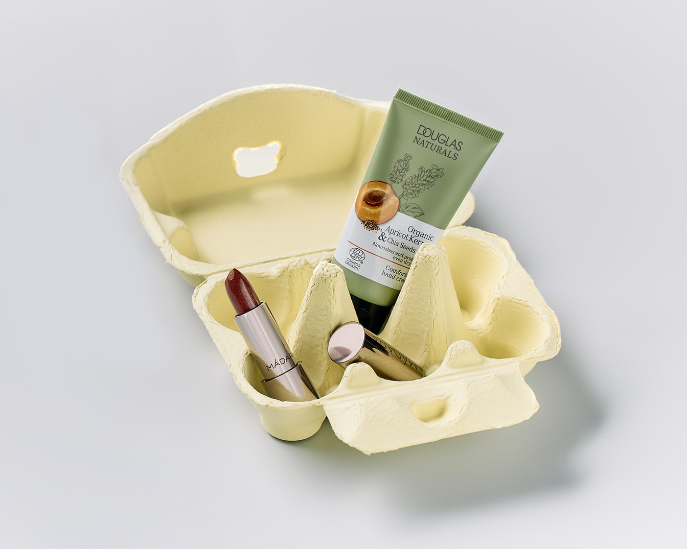 Ecco un vassoio di cartone per uova che contiene un rossetto color prugna di MÁDAF e un tubetto verde di crema mani Douglas Naturals con nocciolo di albicocca e semi di chia.