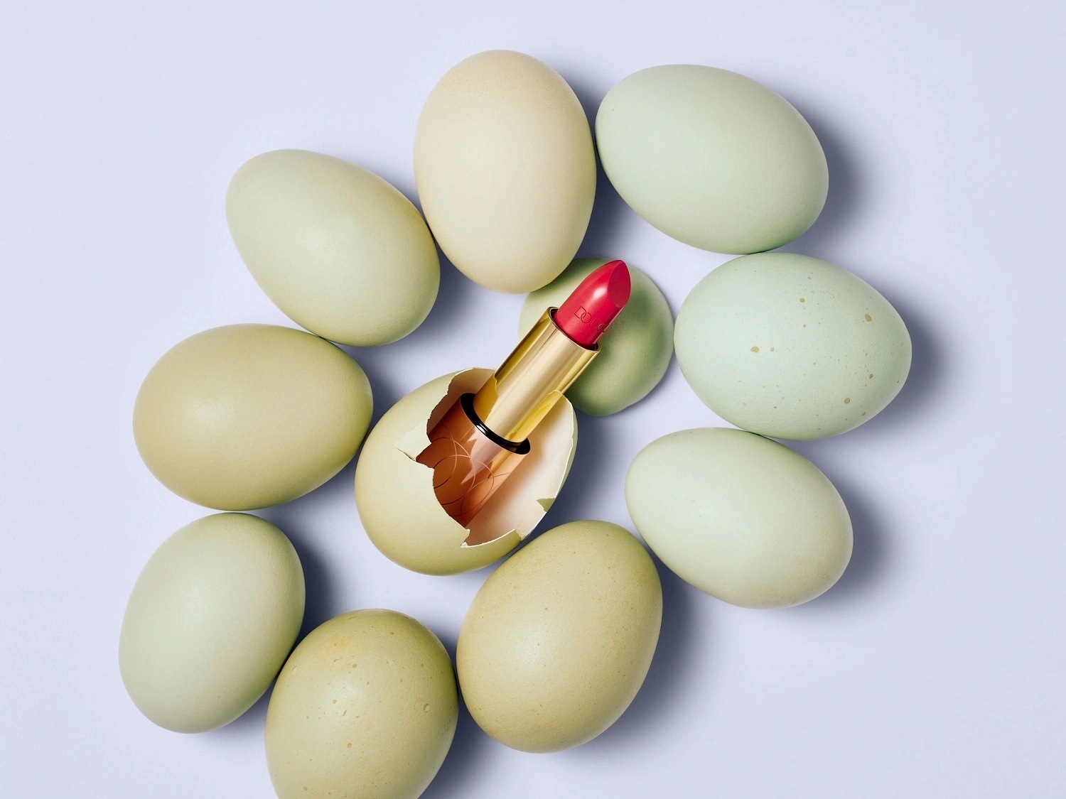 Un rossetto rosso con confezione dorata emerge da un uovo rotto, circondato da uova color pastello su sfondo chiaro.