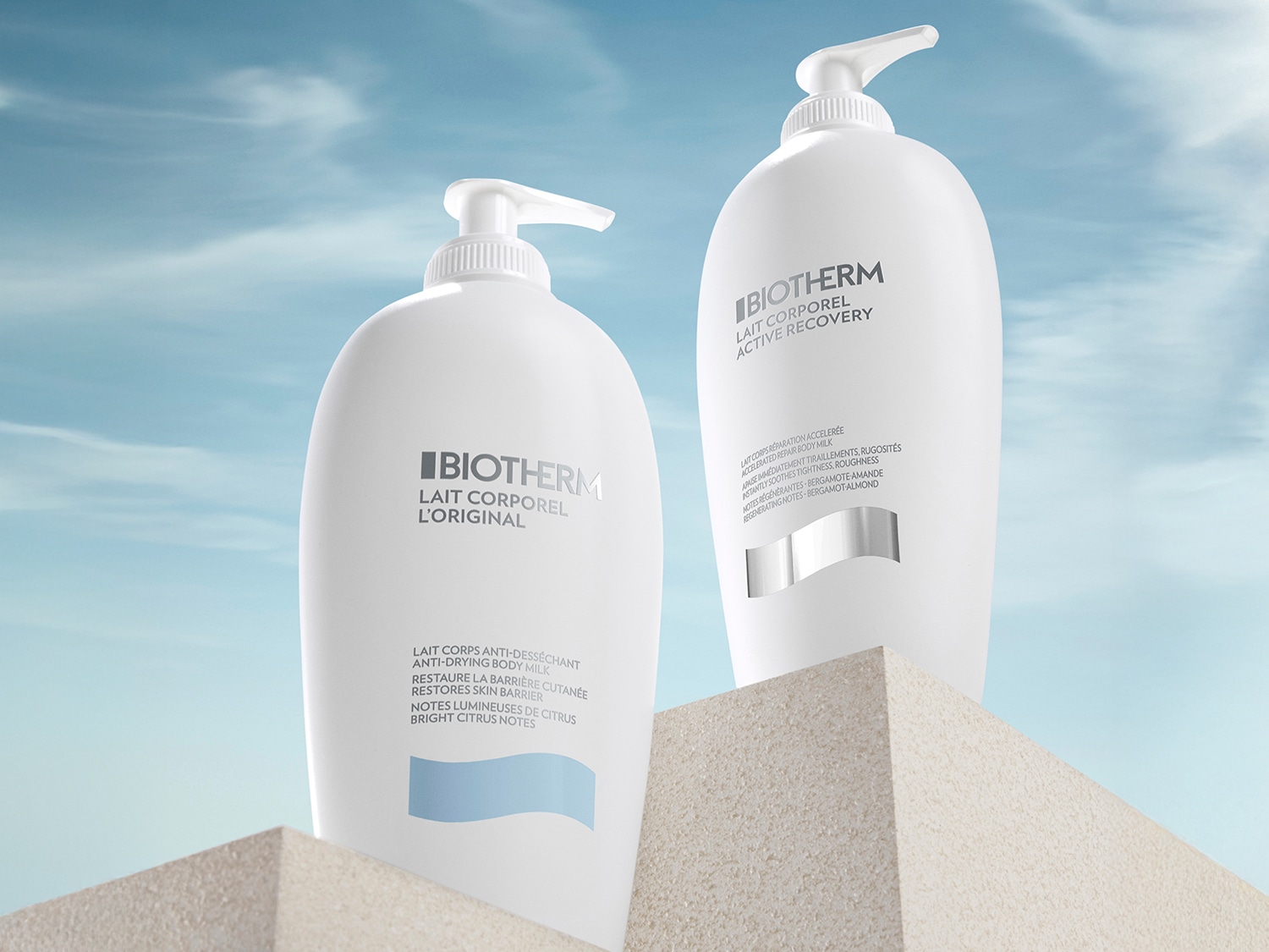 Dos botellas blancas de Biotherm Lait Corporel, una L'Original y otra Active Recovery, sobre una superficie texturizada con un cielo azul claro de fondo.