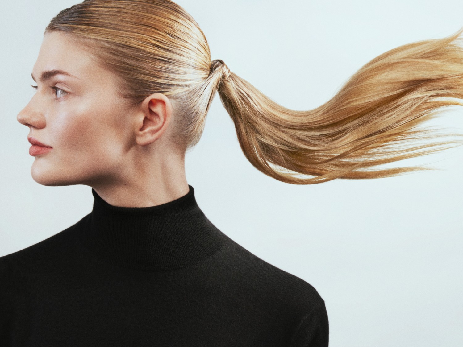 Profiel van een blonde vrouw met een strakke paardenstaart en een zwarte coltrui, haar haar golft zachtjes in de wind.