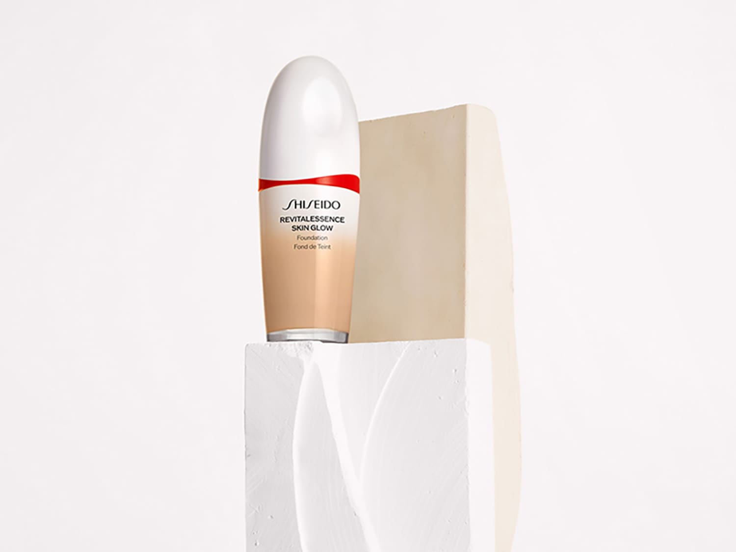 Base de maquillaje Shiseido Revitalessence Skin Glow en un envase degradado de blanco a beige, sobre un pedestal blanco.