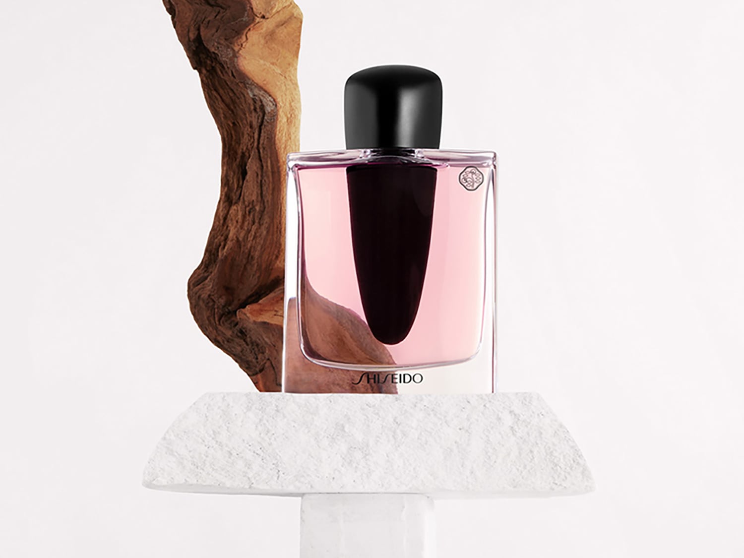 El frasco de perfume Shiseido, con su líquido rosado y tapón negro, se exhibe sobre una base de piedra blanca, acompañado de una pieza de madera rústica.