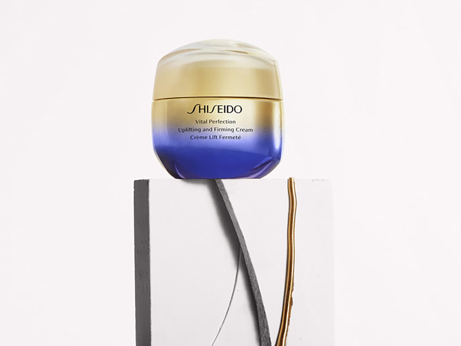 El tarro de crema Vital Perfection Uplifting and Firming de Shiseido tiene una tapa transparente, un cuerpo dorado que se desvanece en azul y está sobre un pedestal blanco.