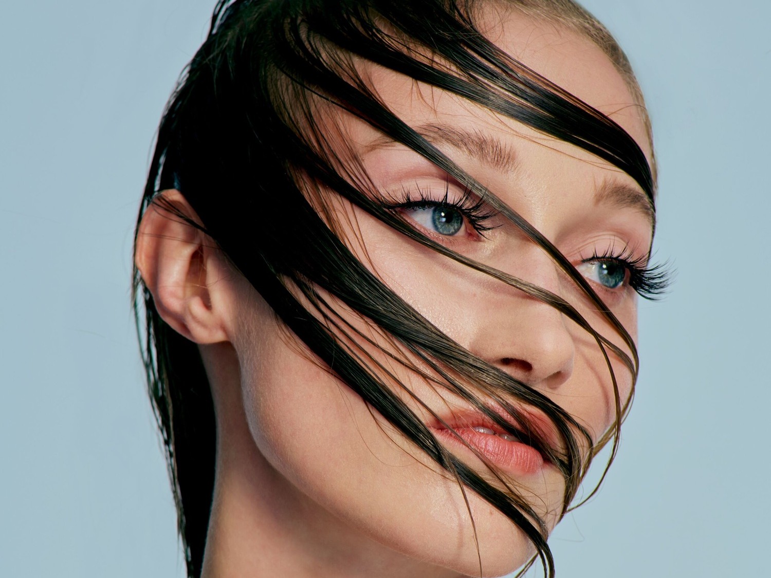 Close-up van een vrouw met nat haar dat over haar gezicht valt, haar blauwe ogen en volle wimpers benadrukken haar natuurlijke schoonheid.