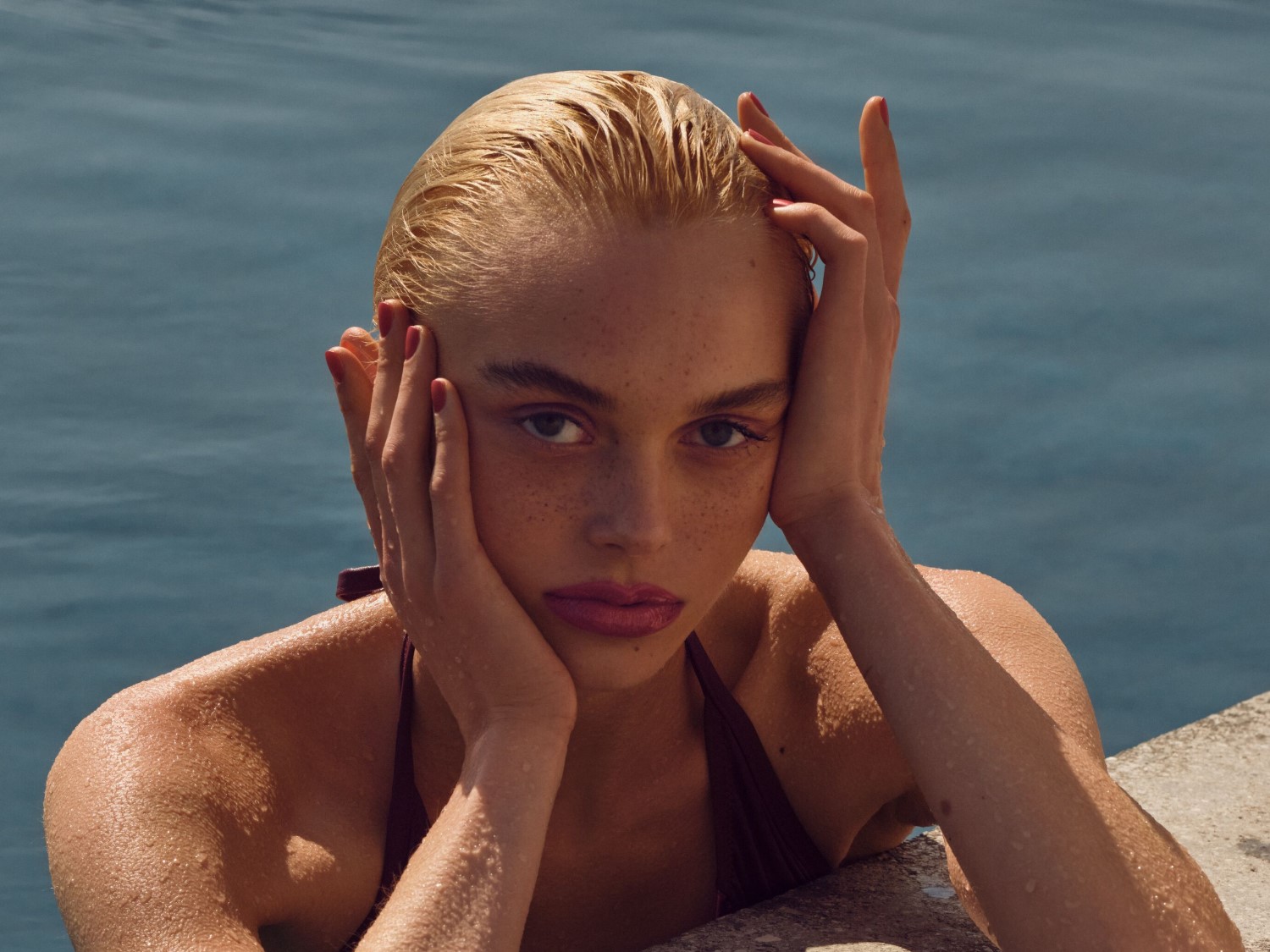 Een vrouw met blond haar en sproeten, leunend op haar handen bij een zwembad, kijkt recht in de camera met een intense blik.