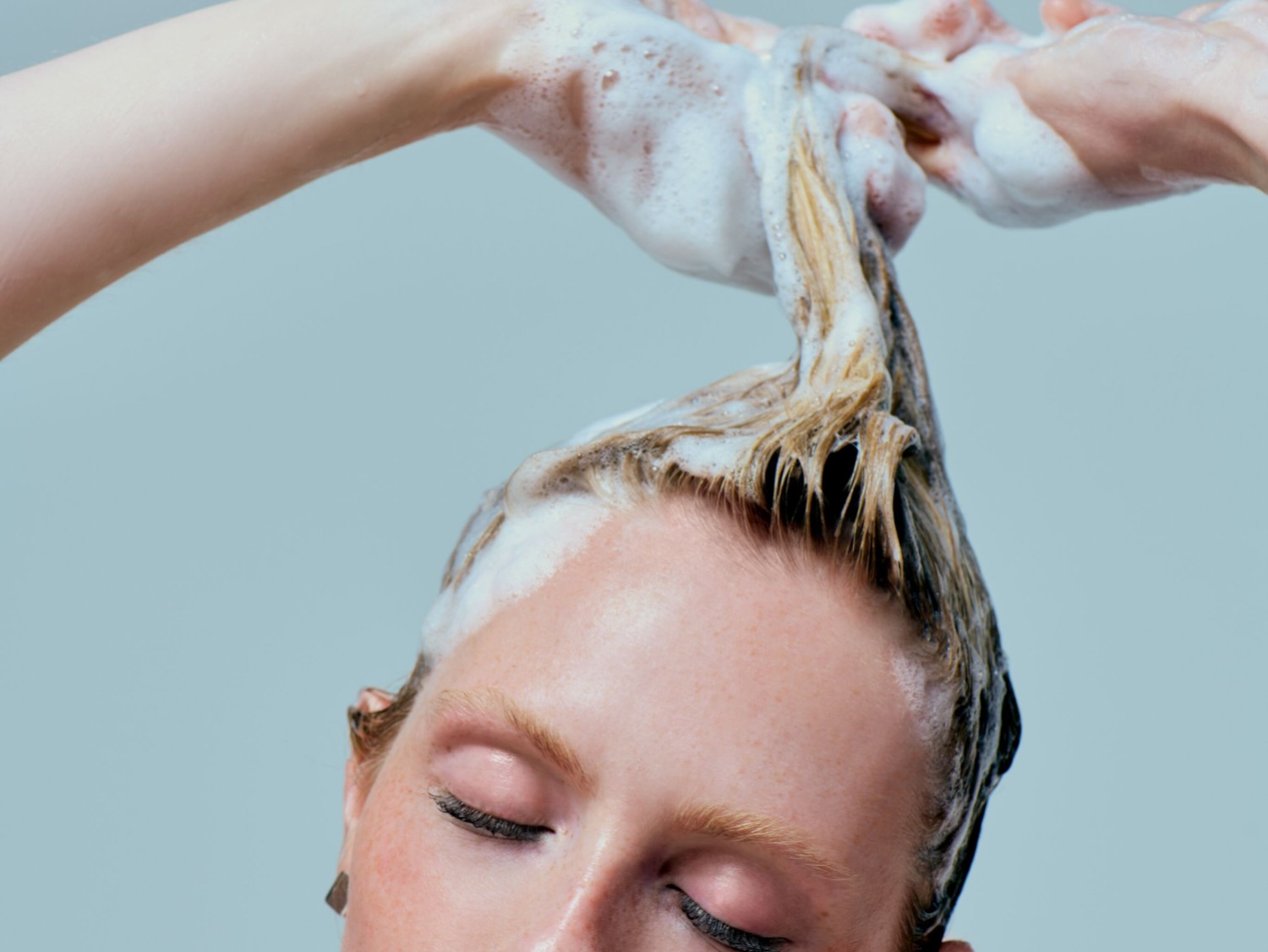 Een vrouw met gesloten ogen wast haar blonde haar met schuimende shampoo.