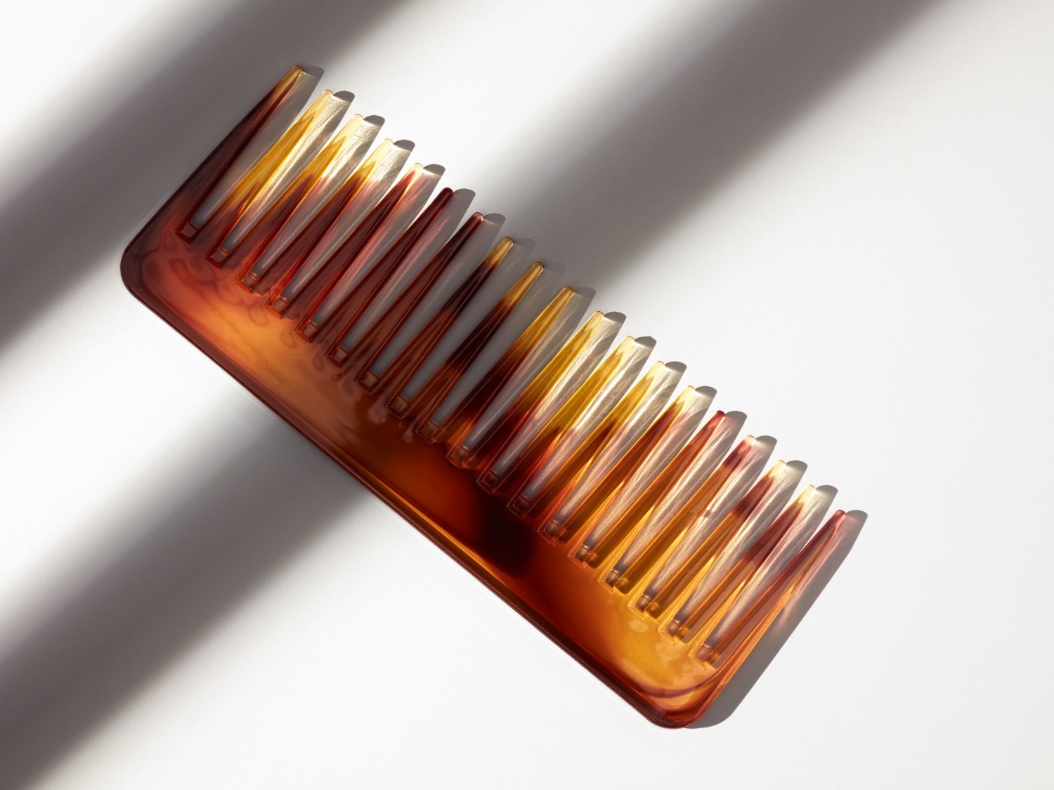 Een rechthoekige haarkam met brede tanden in een mix van amberkleuren, van lichtgeel tot diep bruin, ligt in helder zonlicht op een witte achtergrond.