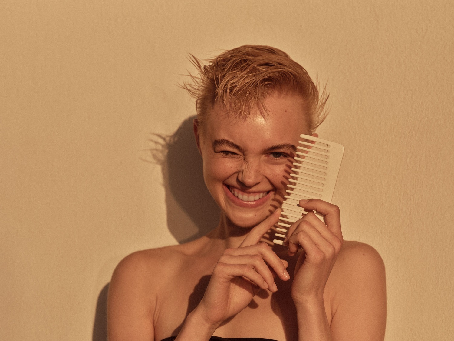 Een lachende vrouw met kort blond haar houdt een witte kam voor haar gezicht, terwijl ze knipoogt en haar tanden laat zien.