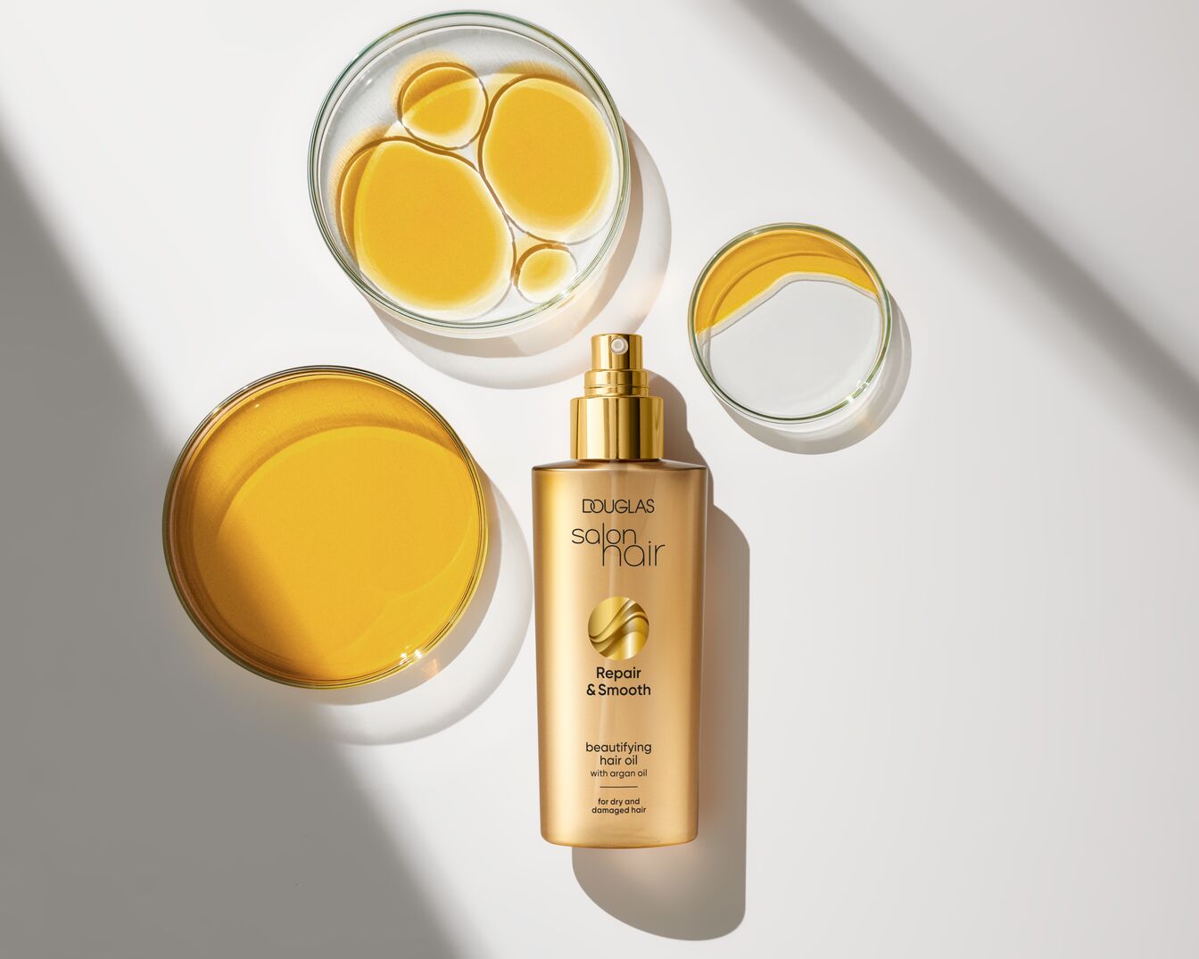 Das Douglas Salon Hair Repair & Smooth Schönheits-Haaröl mit Arganöl in goldener Verpackung, umgeben von Schalen mit goldfarbenem Öl, ideal für trockenes und strapaziertes Haar.