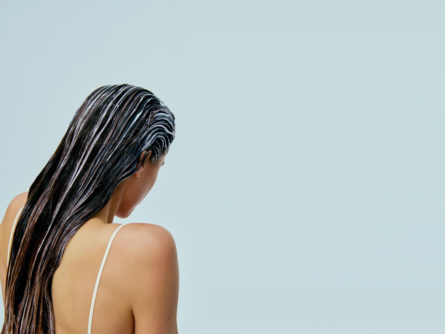 Eine Frau von hinten mit langen, braunen Haaren, die mit einer weißen Creme bedeckt sind, vor einem hellblauen Hintergrund.