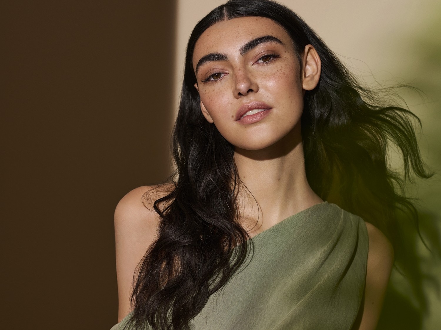 Porträt einer Frau mit langen, gewellten, dunklen Haaren, Sommersprossen und einem grünen Oberteil vor einem hellen Hintergrund.