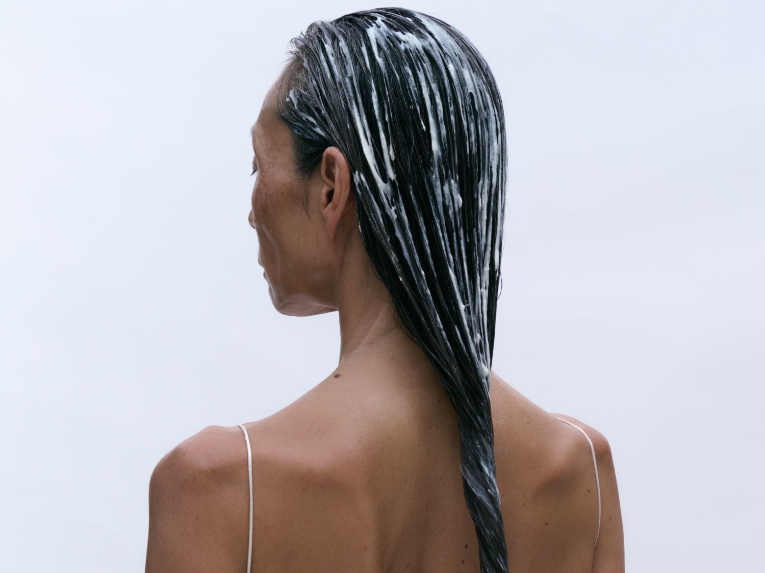 Rückenansicht einer Frau mit dunklem, nassem Haar, das mit einer weißen, cremigen Substanz bedeckt ist, die wie eine intensive Haarbehandlung aussieht.