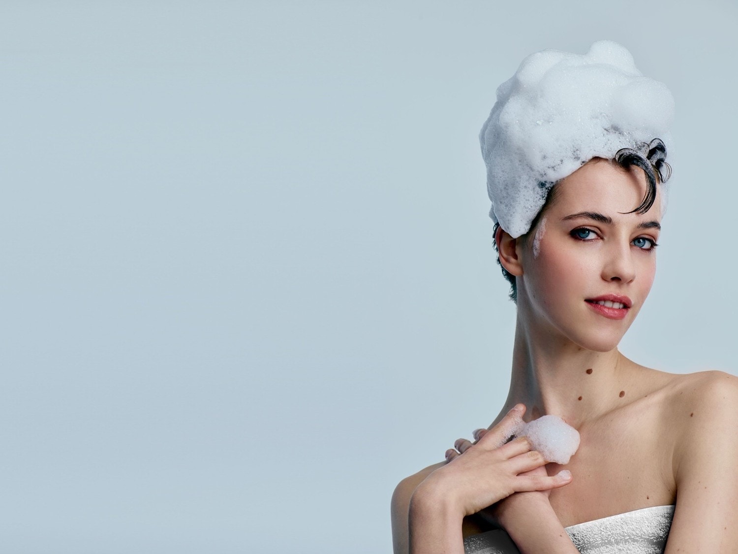 Eine Frau mit kurzen, dunklen Haaren und blauen Augen hat Schaum auf dem Kopf und berührt ihre Schulter mit einer Hand voller Schaum.