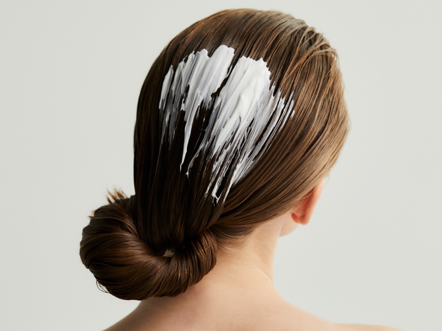 Nahaufnahme von der Rückansicht einer Frau mit braunen Haaren, auf die eine dicke, weiße Haarmaske aufgetragen wird, während sie zu einem Knoten zusammengesteckt sind.