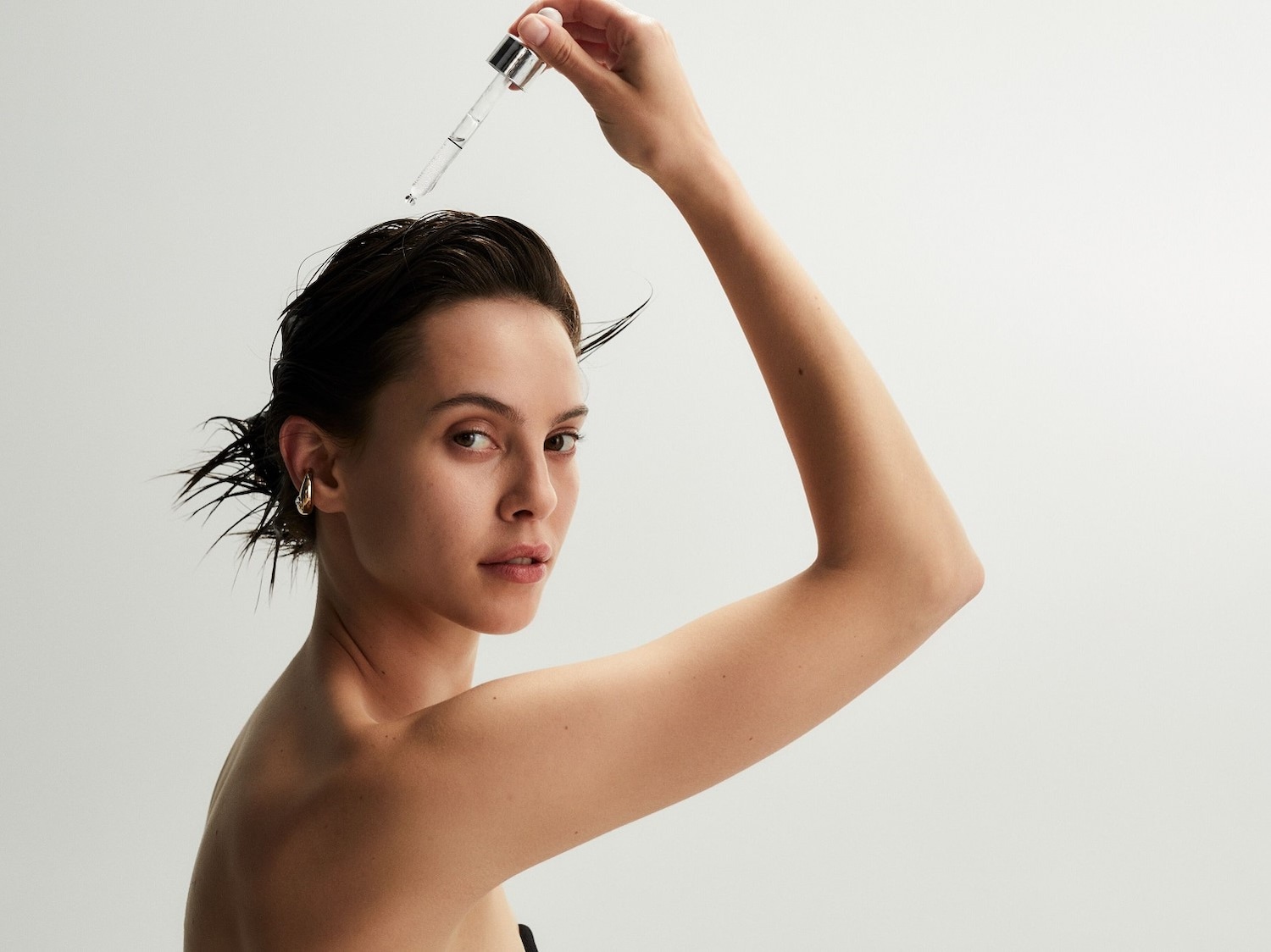 Eine Frau mit nassen, dunklen Haaren, die ein Serum mit einer Pipette auf ihr Haar aufträgt, während sie in die Kamera schaut.