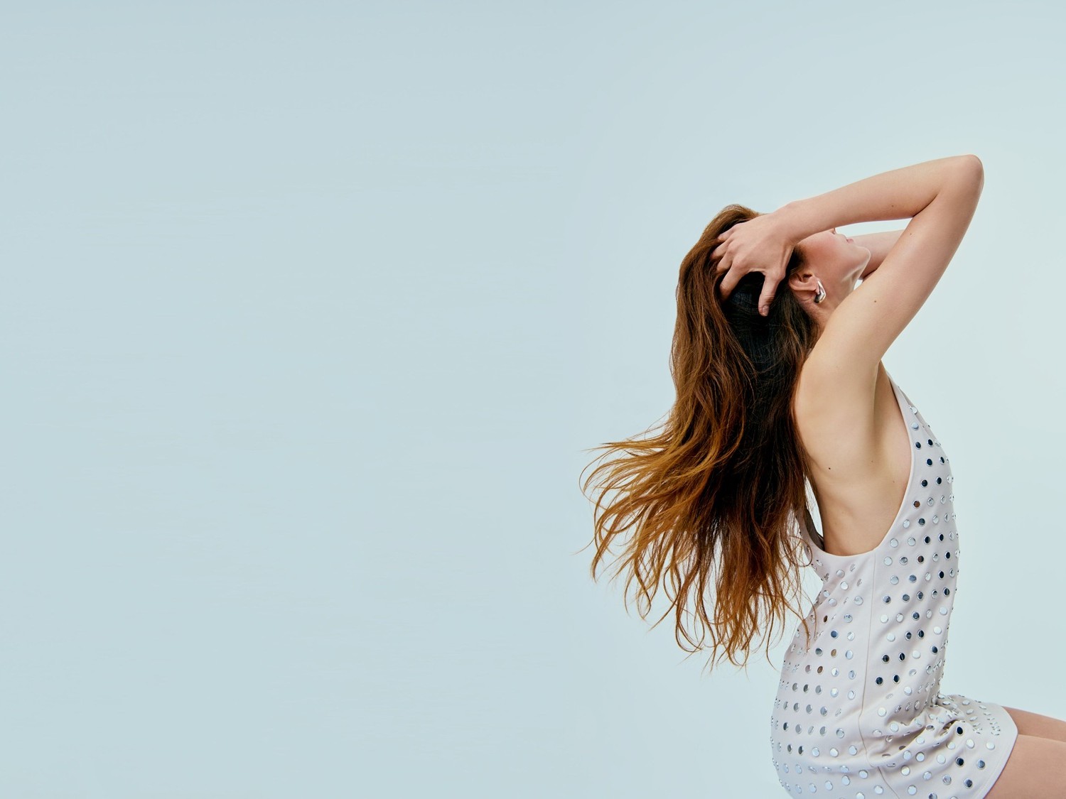 Eine Frau mit gewelltem, braunem Haar, die ein ärmelloses, kurzes Kleid mit silbernen Pailletten trägt, posiert mit erhobenen Armen und nach hinten geneigtem Kopf.