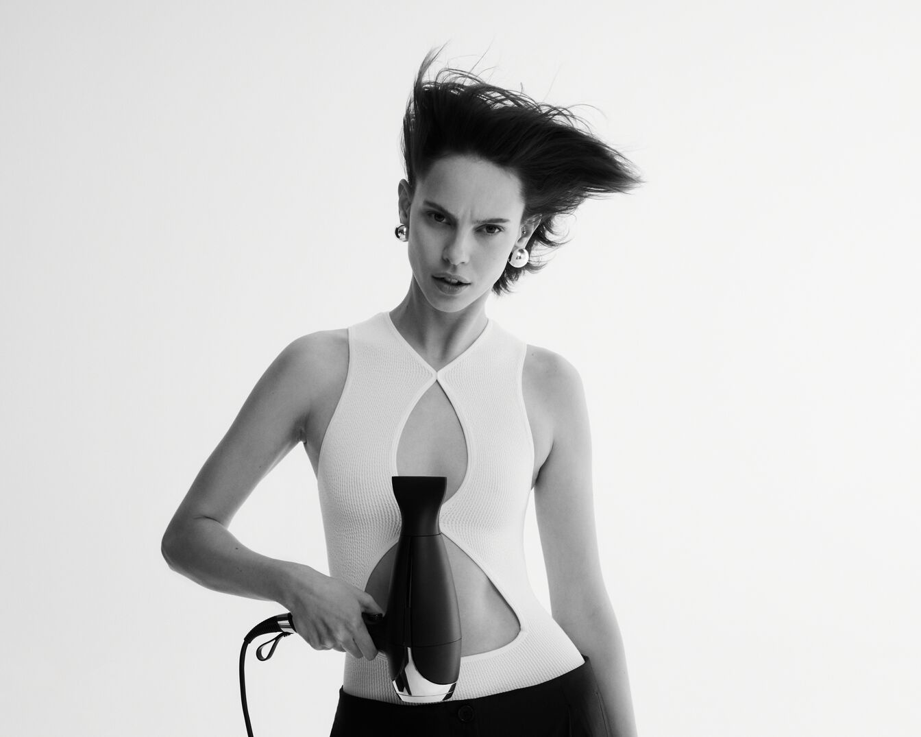 Een zwart-wit foto van een vrouw met een wit topje en zwarte broek, die een zwarte föhn vasthoudt terwijl haar haar opwaait.