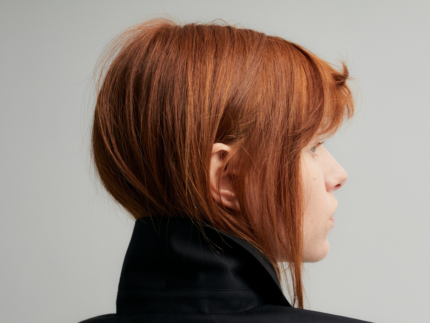 Profil einer Frau mit kurzen, roten Haaren und einem schwarzen Oberteil vor einem neutralen Hintergrund.